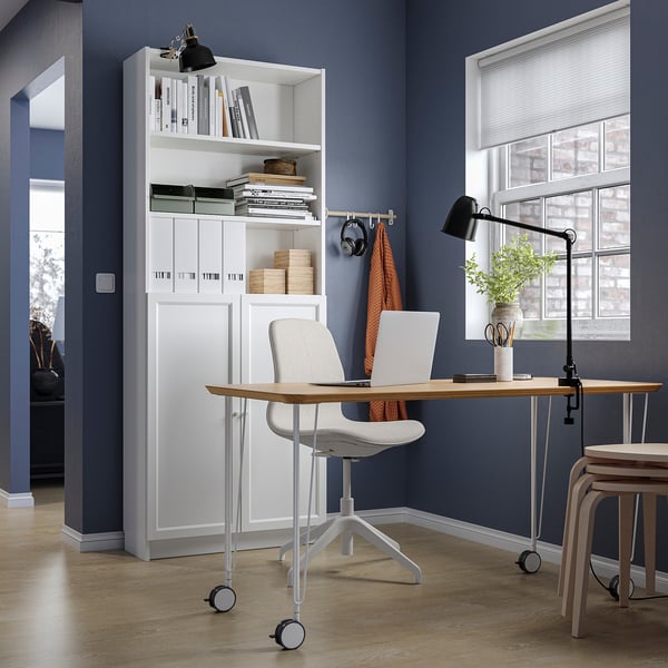 Modern home office with white BILLY bookcase, lightwood desk, grey chair, black lamp, and window.