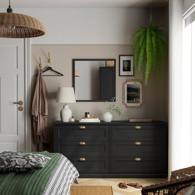 Stylish bedroom with black oak FÅGELFJÄLLET dresser, mirror, and hanging plant. White lamp, decor, and hanging coat, with a bed partially visible.