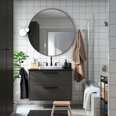 Modern bathroom with dark grey HAVBÄCK vanity, round mirror, white sink, black fixtures, wooden stool, large plant, and towels.