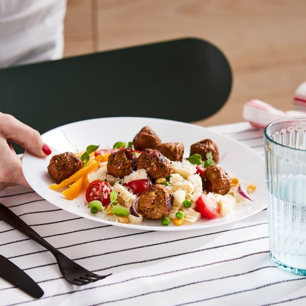 A person holds a plate of vegetable balls with colourful vegetables. A glass and cutlery are on a striped tablecloth.