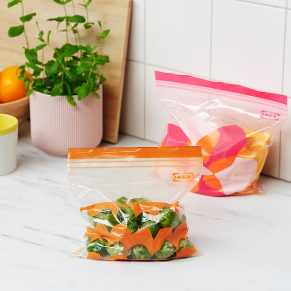 Two IKEA zip bags, one holding greens, displayed on a countertop with plant and fruit.