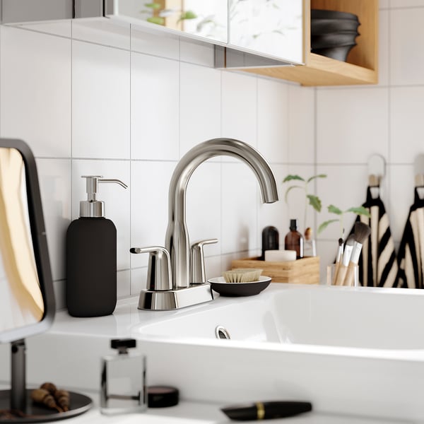 Modern bathroom tap, MACKSJÖN, in metallic finish, with smooth lines on white countertop. Durable stainless-steel, surrounded by toiletries.