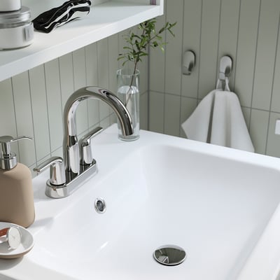A modern bathroom sink with chrome tap, holding a small plant and soap dispenser, attached to a wall with light green vertical tiles.