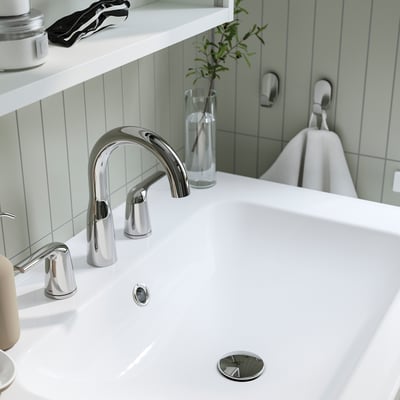 A modern bathroom sink with a chrome tap and pop-up drain, set against light green tiles.