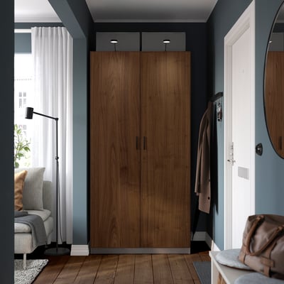 A tall, dark wooden PAX wardrobe. It has two sections, with hooks for coats and a bag placed nearby. A grey couch and a lamp are visible on the left.