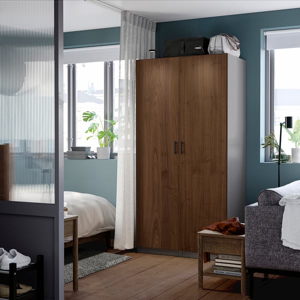 Modern bedroom with tall wooden PAX wardrobe, grey armchair, bed, and plants near windows.