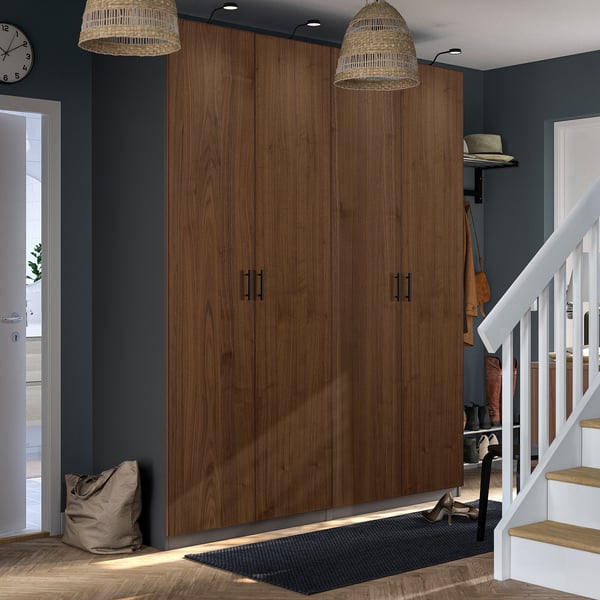 Large brown wooden PAX closet against dark wall, flanked by wicker lamps, near white staircase and black doormat.