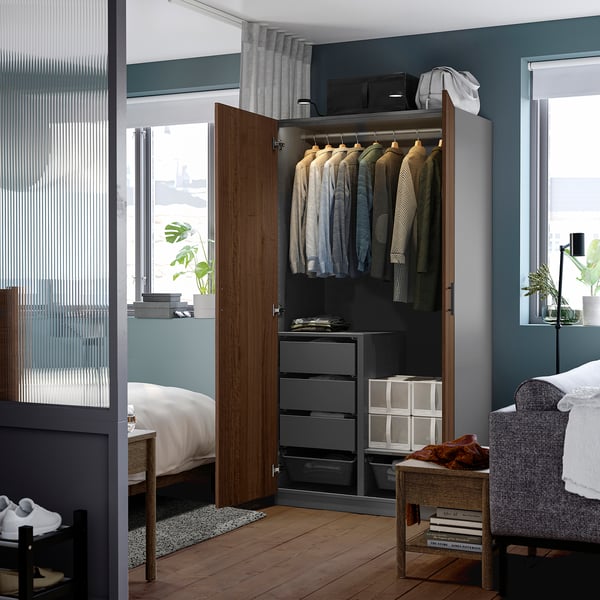 Modern bedroom showcasing an open PAX wardrobe with shirts, drawers, and storage boxes; adjacent window and plant.