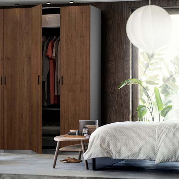 Modern bedroom with open wooden closet displaying clothes, featuring PAX system. Soft light, plant, and cosy bed with white bedspread.
