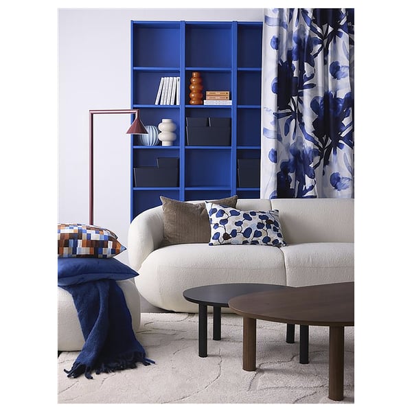 Modern living room with white sofa, blue pillows, wooden tables, blue bookshelf, and floral curtain.