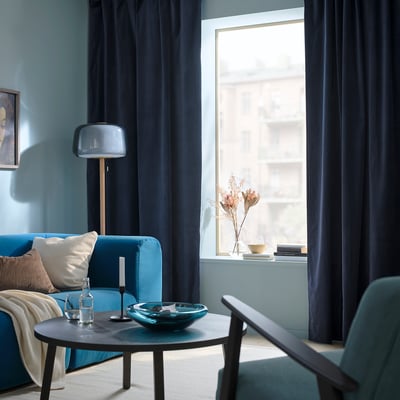Modern living room with blue curtains, teal sofa, black coffee table, and tall lamp by window with dried flowers.