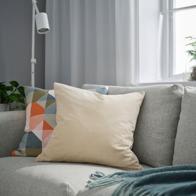 Living room with SANELA beige cushion, multicoloured pillow, green throw, plant, lamp, and window with curtain.