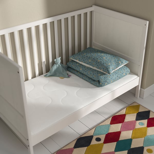 Small white toddler bed with firm mattress and blue patterned blanket, cuddly toy, and multicoloured rug on the floor.