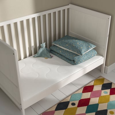 Small white toddler bed with firm mattress and blue patterned blanket, cuddly toy, and multicoloured rug on the floor.