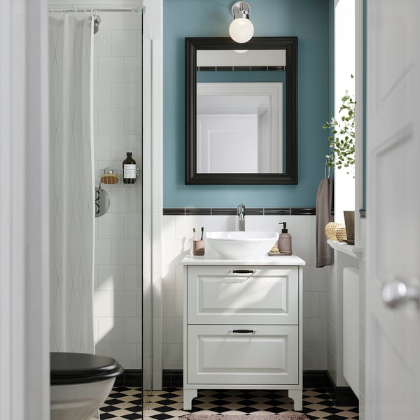 White vanity with sink, 2 drawers, mirror, shower, green plant.