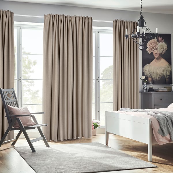 Bedroom with ANNAKAJSA room-darkening beige curtains, a white bed, grey chair, and floral artwork. Curtains reach floor, framing large window.
