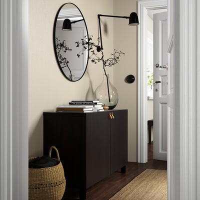 Modern living room with BESTÅ grey sideboard, oval mirror, black lamp. Sideboard holds books and vase. Nearby basket and minimalist door.