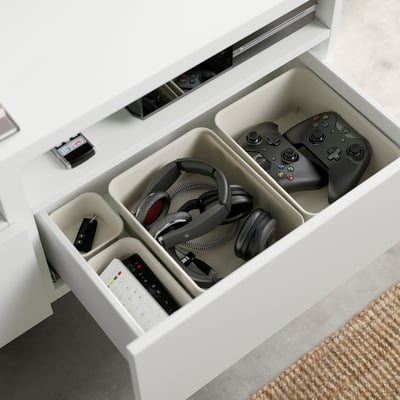 White BESTÅ unit with open drawer showing organised gaming and tech accessories including headphones, game controllers, and remotes in compartments.