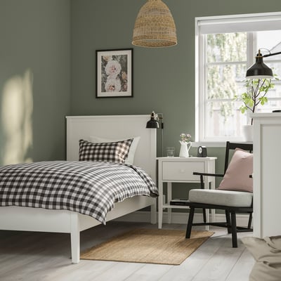 White bed, chequered bedding, side table, lamp, grey chair, pink cushion, wicker light, floral art, rug, plant.