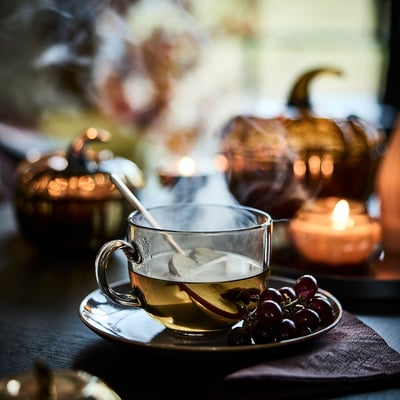 A steaming cup of tea on a saucer with grapes beside it, part of an autumn-themed table setting.