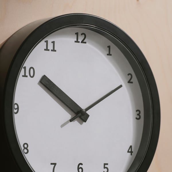Black wall clock with thick hour hand pointing at 10 and minute hand near 45, in modern minimalist design, mounted on a beige wall.