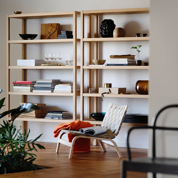 Wooden bookshelf STOCKHOLM 2025 with books, vases, and a plant. White chair with orange fabric in front.