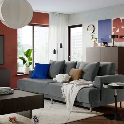 Modern living room: grey sofa, colourful pillows, red wall, large window, rug, toys, plants, art.
