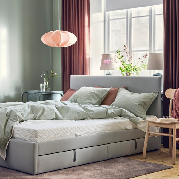 Modern bedroom with gray ÅBYGDA mattress, light green pillows, wooden side table, pendant lamp, and large window with red curtains.
