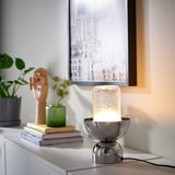 Modern silver ACKJA table lamp with textured glass & metallic base on cabinet, surrounded by small plants, books, and a decorative sculpture.
