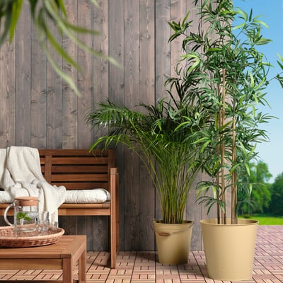 Two ÅKERBÄR beige pots hold large green plants, placed beside a brown wooden bench with white cushions on a tiled patio, against a wooden wall.
