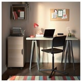 Modern home office with beige ALEX storage, wooden desk, black chair, red lamp, laptop, and colorful rug.
