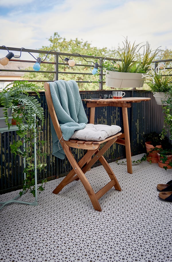 Cozy balcony setup with an ALTAPPEN white plastic floor deck, wooden folding chair, cushions, table, hanging lights, and potted plants.