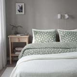 Neat bedroom with modern design: grey-green patterned bedding, wooden nightstand with books, glass, and plant. Two adjustable silver lamps beside bed.