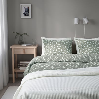 Neat bedroom with modern design: grey-green patterned bedding, wooden nightstand with books, glass, and plant. Two adjustable silver lamps beside bed.