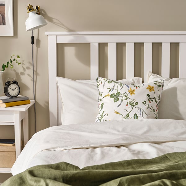 Cozy bedroom with white bed, green blanket, floral pillow. Wall clock left, white wall lamp.
