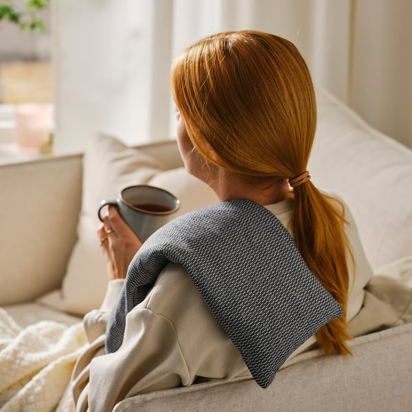 A woman with red hair sits on a beige couch, drinking from a grey mug. She has a grey and white heating pad wrapped around her shoulder.