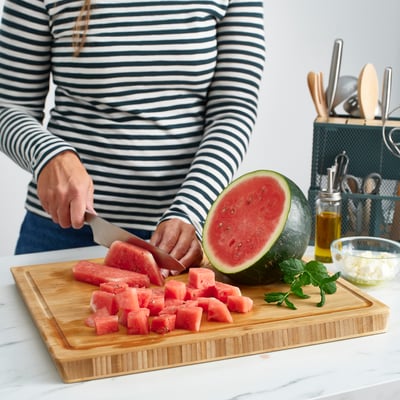 An ikea APTITLIG chopping board, light brown, made of bamboo with a groove on one side and slanted edges for easy grip.