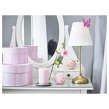 A tabletop lamp with a white shade sits near a mirror and pink boxes. It provides soft, diffused light.