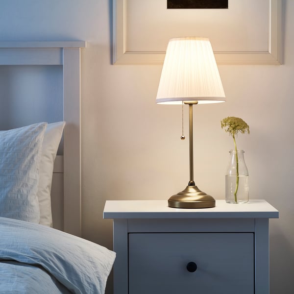 A small, elegant ÅRSTID bedside lamp with a white shade and brass finish base on a blue drawer unit next to a bed.