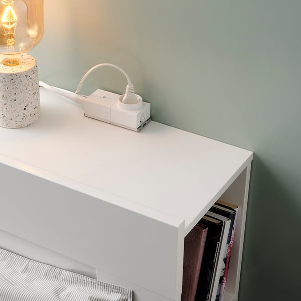 An a white surface with an ÅSKVÄDER power module featuring an on/off switch and a bookshelf containing books, against a light green wall.