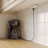 Black coffee maker on wooden counter above white-tiled wall, connected to ÅSKVÄDER power module with cord.