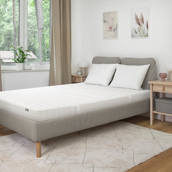 Modern minimalist bedroom with white mattress, gray bed frame, wooden floors, rug, nightstand, lamp, two wall pictures, neat bed, large window.