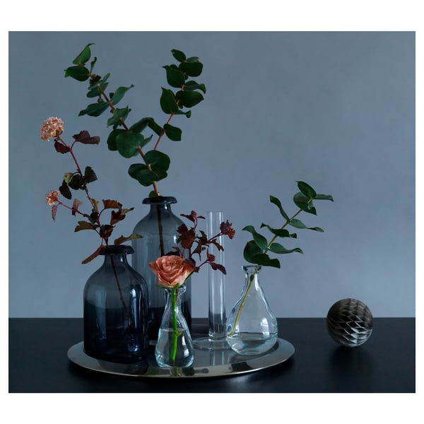 Simple still life with ÅTGÅNG blue, clear glass vases holding flowers on metal tray, gray background.