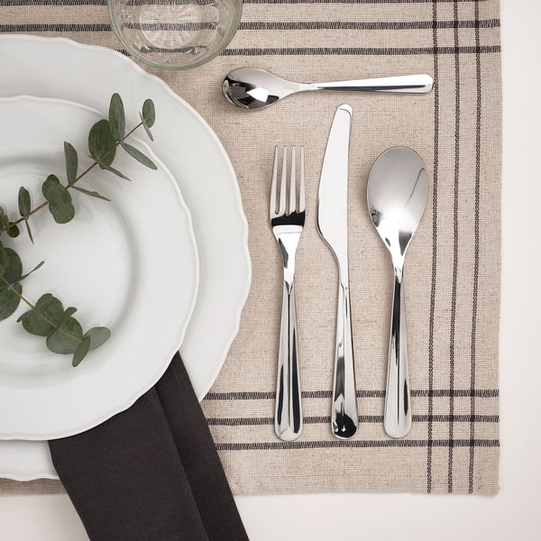 BÄCKÖRING stainless steel cutlery set: fork, knife, teaspoon, and soup spoon on striped placemat next to white plate with greenery.