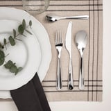 BÄCKÖRING stainless steel cutlery set: fork, knife, teaspoon, and soup spoon on striped placemat next to white plate with greenery.
