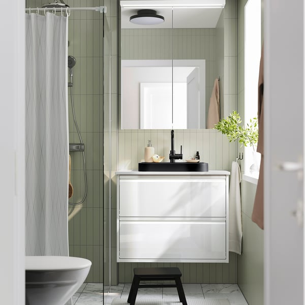 Modern bathroom with semi-recessed black marble basin, white cabinet, green tiles, glass shower, towel hooks, and plant.