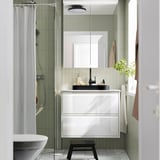 Modern bathroom with semi-recessed black marble basin, white cabinet, green tiles, glass shower, towel hooks, and plant.