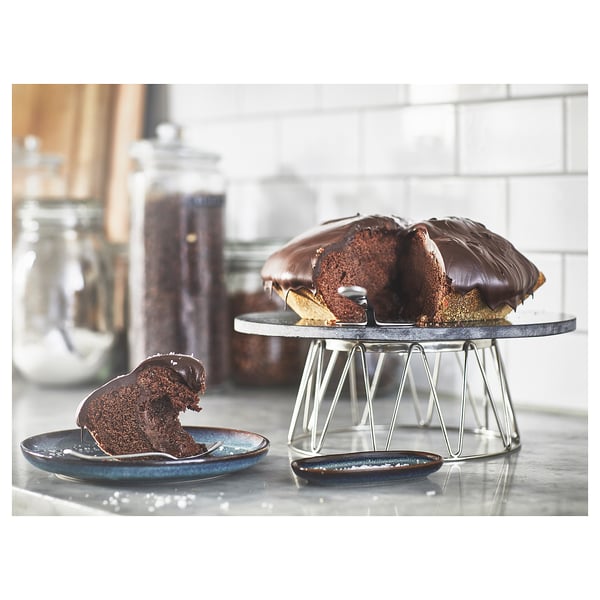 A chocolate cake on a metallic, frosted cake stand. Slice served on a small, blue plate beside it.