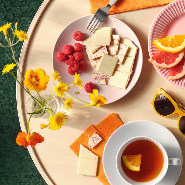 Plate with white chocolate, raspberries, and a cup of tea. Flowers and citrus slices nearby.