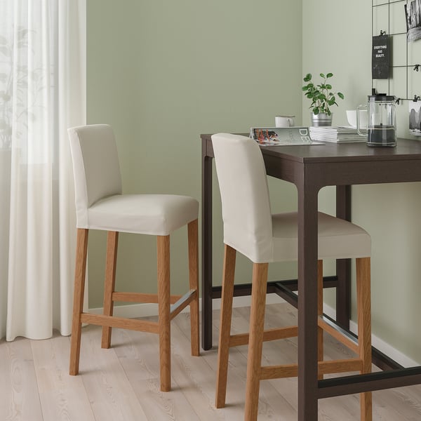 Two BERGMUND bar stools in beige and light wood at dark counter, with cushioned seats and backs, set against light green wall and light wooden floor.